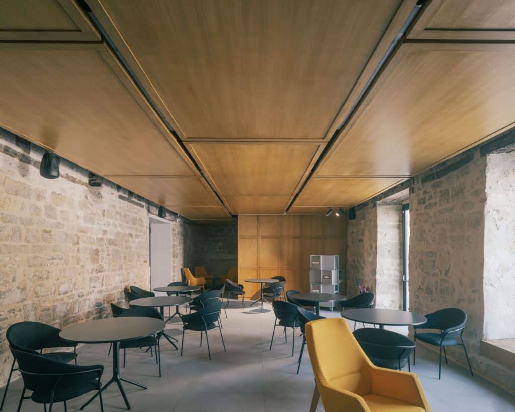 Modern cafeteria inside La Manufacture Cultural Center with black tables, yellow chairs, and exposed stone walls.