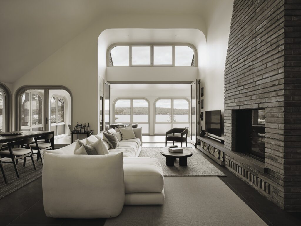Luxurious interior of the Maison de la Pointe living room, showing a white L-shaped sectional sofa and an open wall of arched windows revealing a wide panoramic view of Lake Memphremagog.
