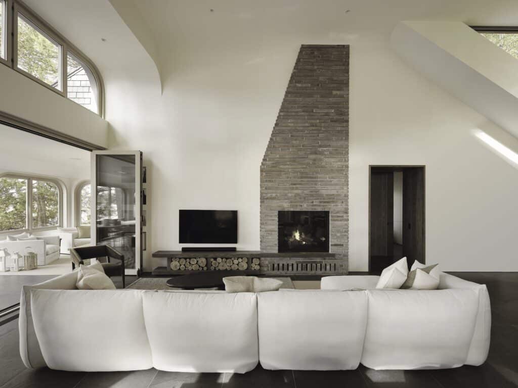 Rear view of the L-shaped white sofa in the Maison de la Pointe living room, facing a large, asymmetrical grey brick fireplace with integrated log storage.