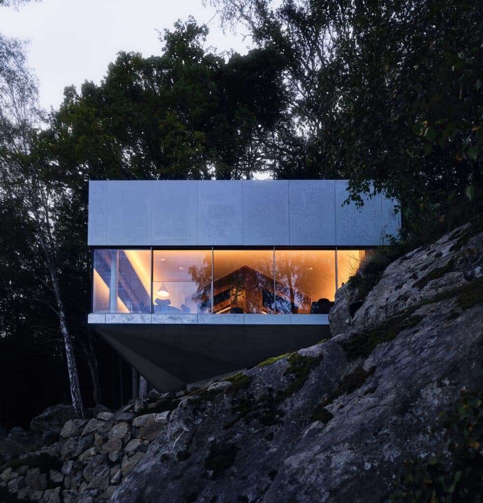 Night view of the main house interior through floor-to-ceiling glass windows, showing the warm wooden central core.