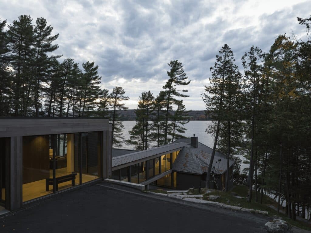 Aerial view of Maison de la Pointe at dusk, showing the new modern wing connected by a fully glazed corridor to the preserved traditional cottage on the lakefront.