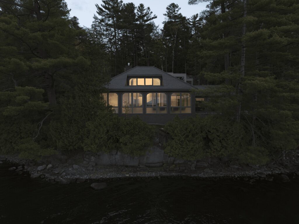 Front view from the water of the Maison de la Pointe traditional cottage at night, its arched windows glowing with warm light on the rocky shore of Lake Memphremagog.