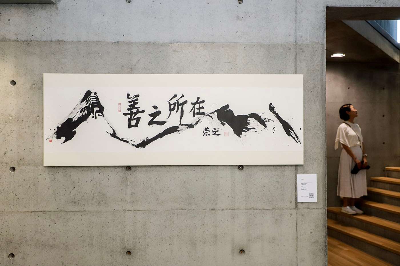 Large ink painting displayed on an exposed concrete wall inside an art gallery with a visitor on wooden stairs.