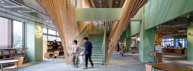 Children and visitors sit inside the Hidamari reading room behind a glass wall with tiered yellow seating at Tsuki no Mori Library
