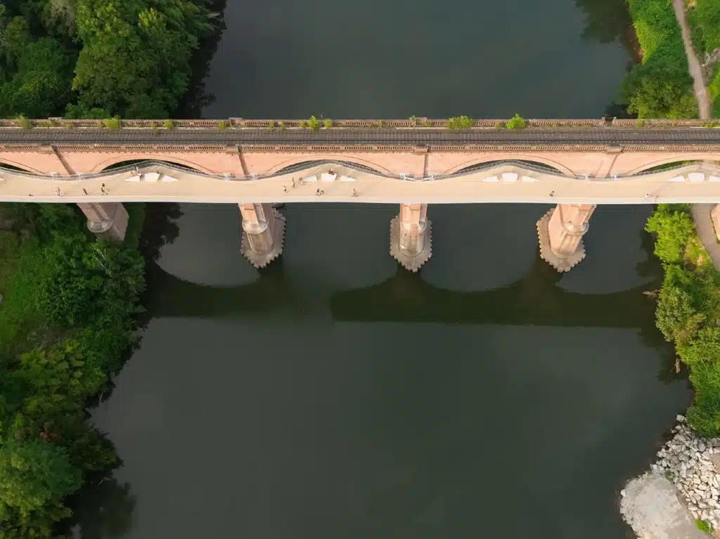 Pedestrians walking and resting on a curved steel footbridge attached to a brick railway viaduct in Albi.