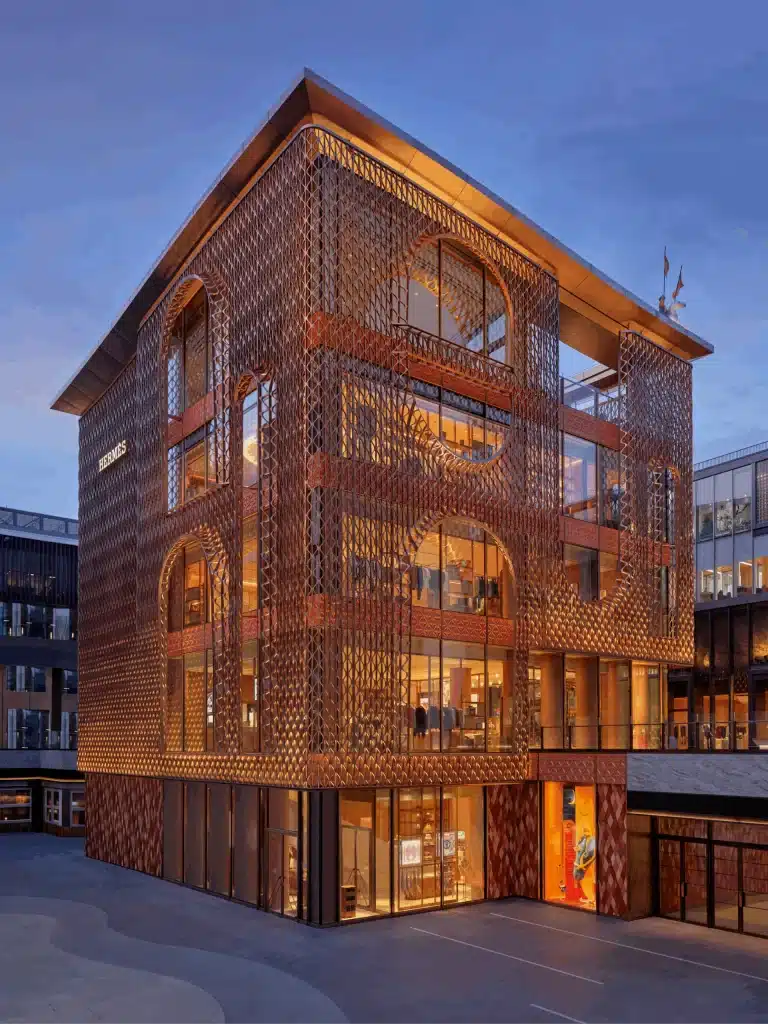 الواجهة السيراميكية لمتجر هيرميس بكين عند الغسق، مع ضوء دافئ يتوهج من الداخل عبر الشاشة.