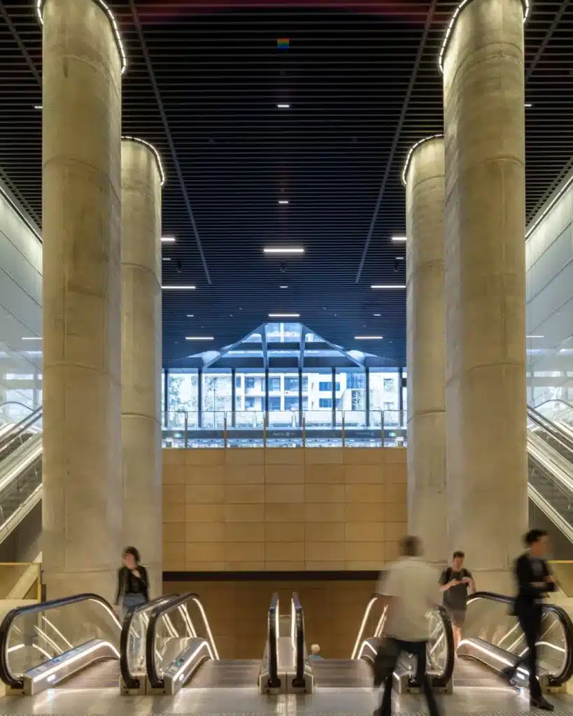 Monumental concrete columns flanking escalators at Gadigal Metro Station Park Street entrance with sandstone wall panels and dark slatted ceiling