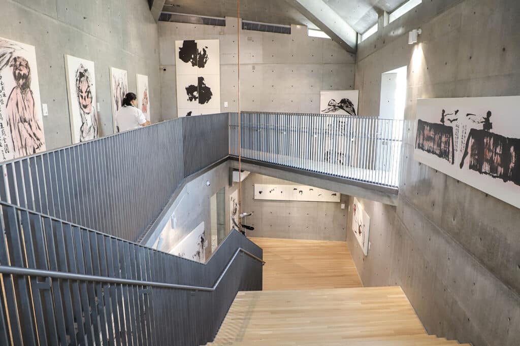Multi-level gallery interior featuring exposed concrete walls, wooden floors, and metal railings with visitors viewing art.