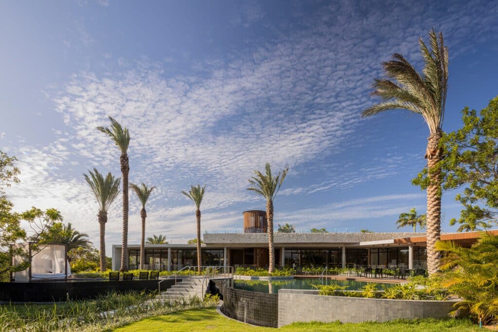 Exterior view of Quintas do Lago Praia dos Coqueiros project showing the organic infinity pool, palm trees, and the horizontal concrete architecture under a blue sky.