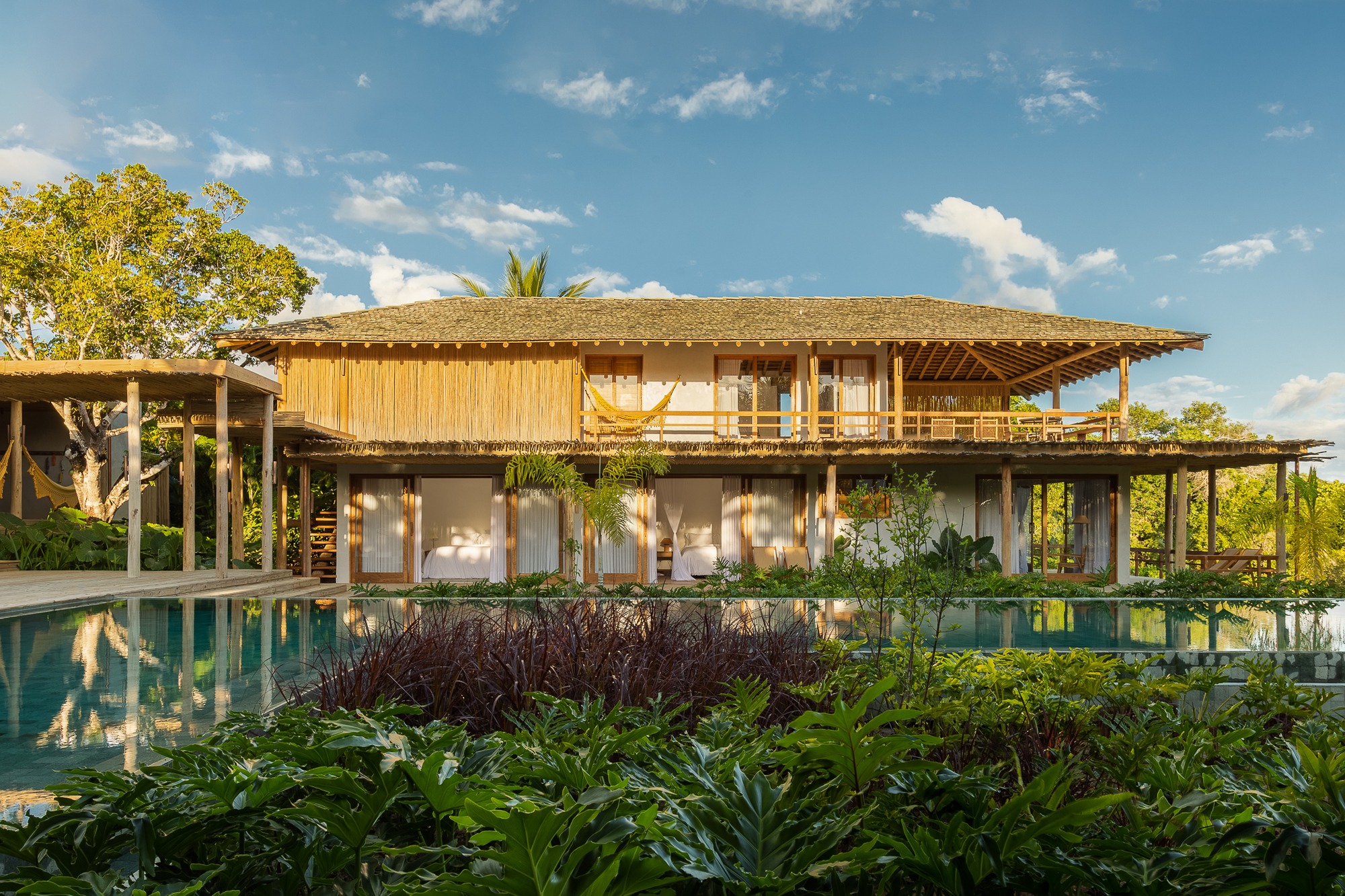 Exterior view of MP House featuring a wooden structure, clay tile roof, and an infinity pool reflecting the tropical landscape.