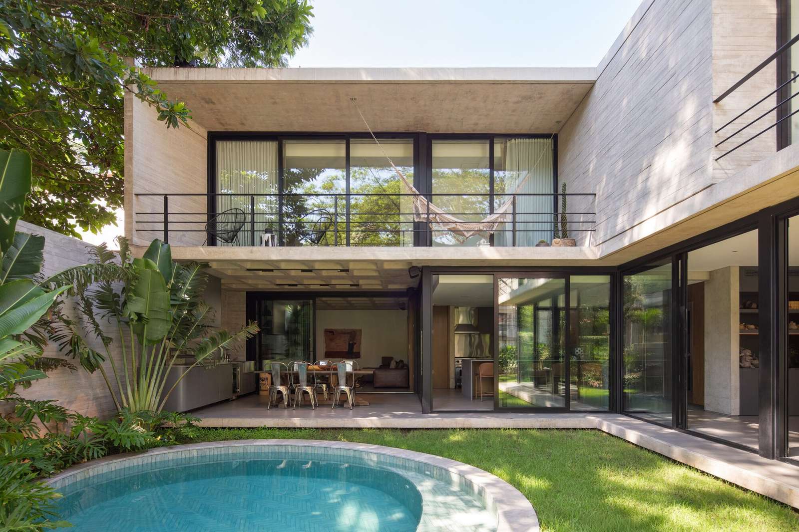 Rear view of Casa Refugio showing the L-shaped concrete structure, glass sliding doors, outdoor swimming pool, and lush garden.