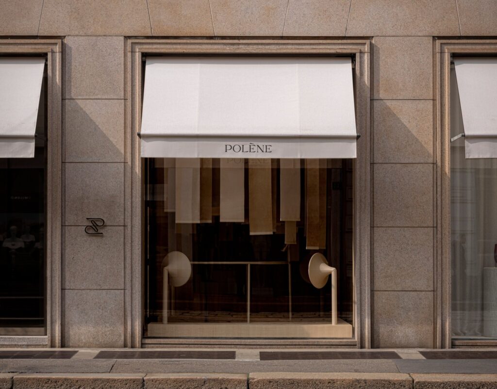 Minimalist storefront of Pol&egrave;ne Milan with stone cladding, white awnings, and a curated window display featuring textured fabric panels.