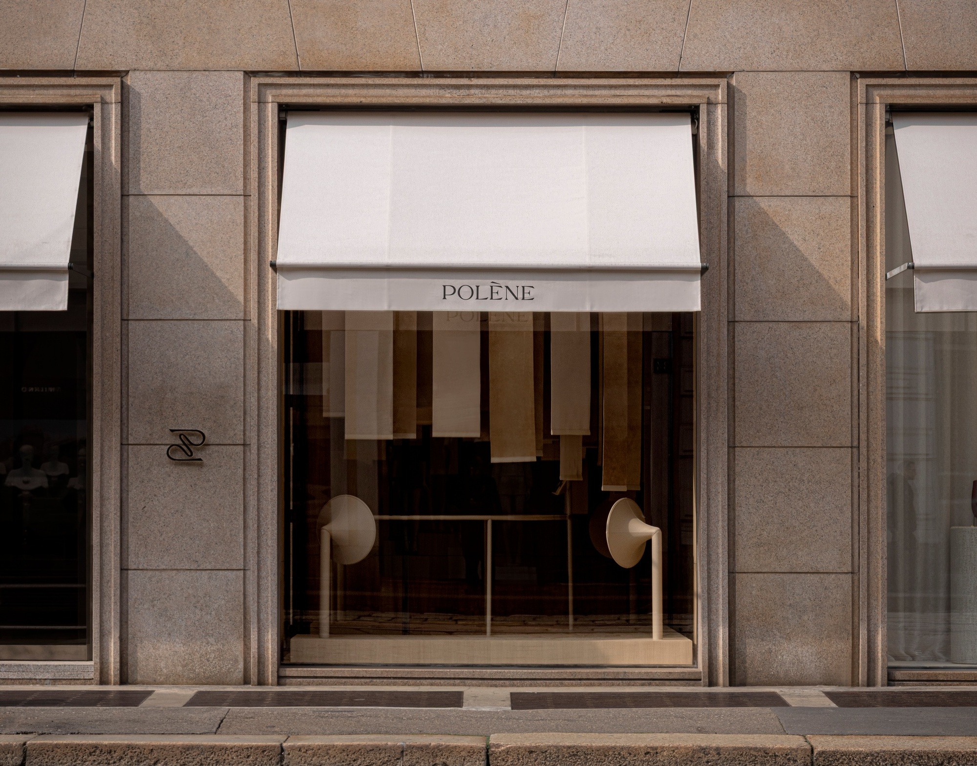 Minimalist storefront of Polène Milan with stone cladding, white awnings, and a curated window display featuring textured fabric panels.