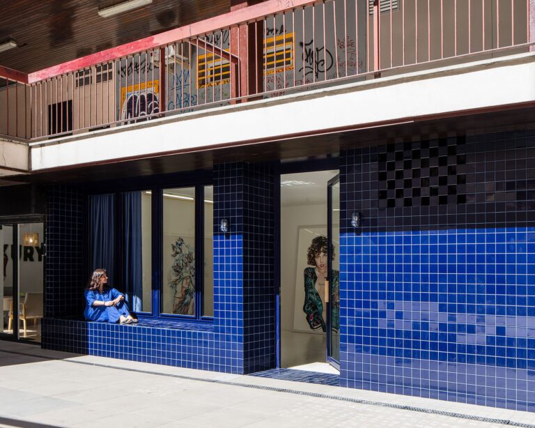A woman sitting on a cobalt blue ceramic tile bench integrated into a Portuguese-style facade under a concrete overhang.