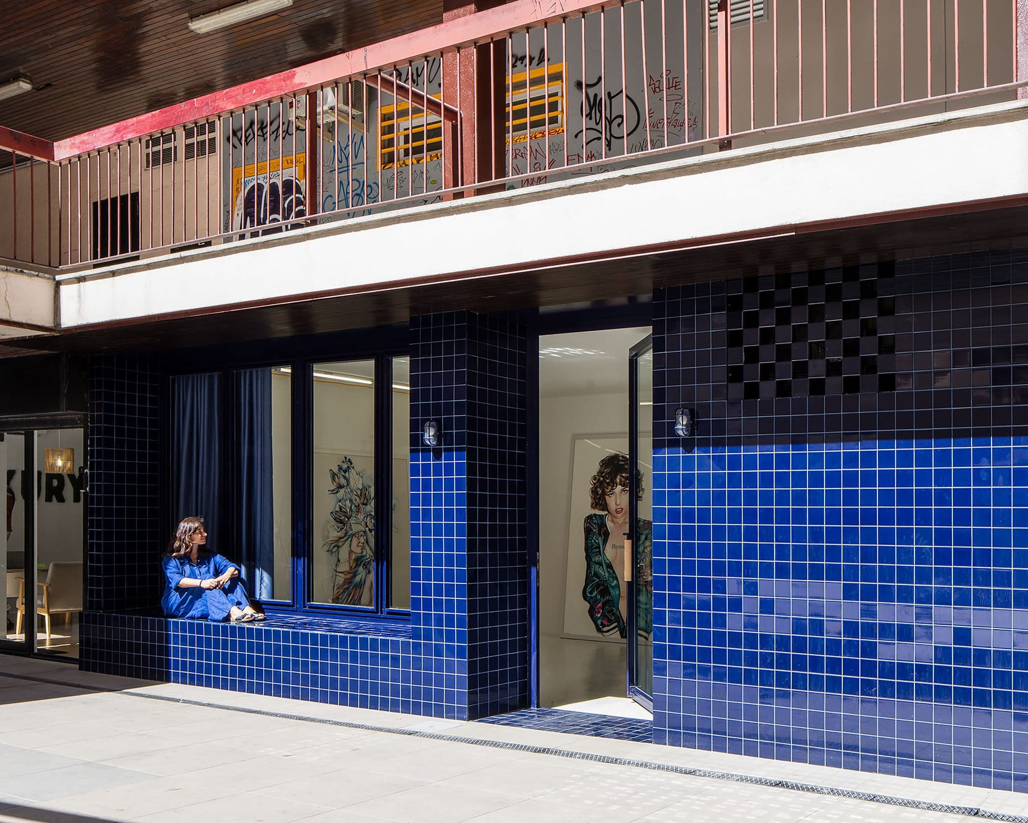 A woman sitting on a cobalt blue ceramic tile bench integrated into a Portuguese-style facade under a concrete overhang.