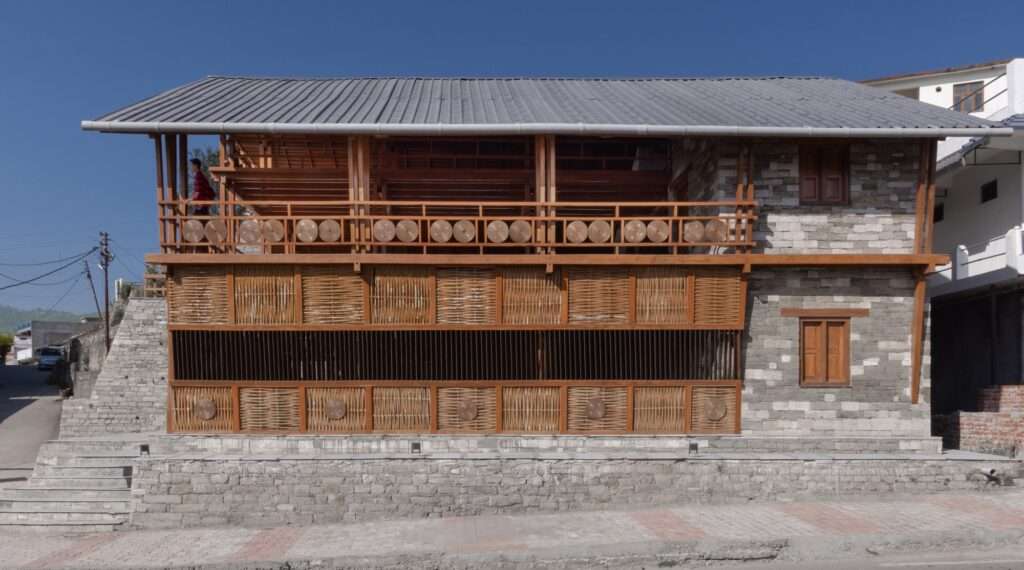 Side elevation of Champawat Market Plaza featuring local stone walls and a modular wooden balcony with woven bamboo screens.