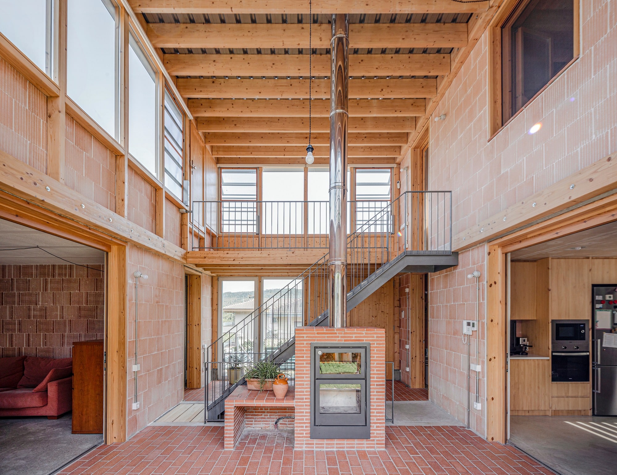 Interior view of a central atrium in a bioclimatic house featuring exposed brick walls, timber ceilings, a central wood stove, and a metal staircase.