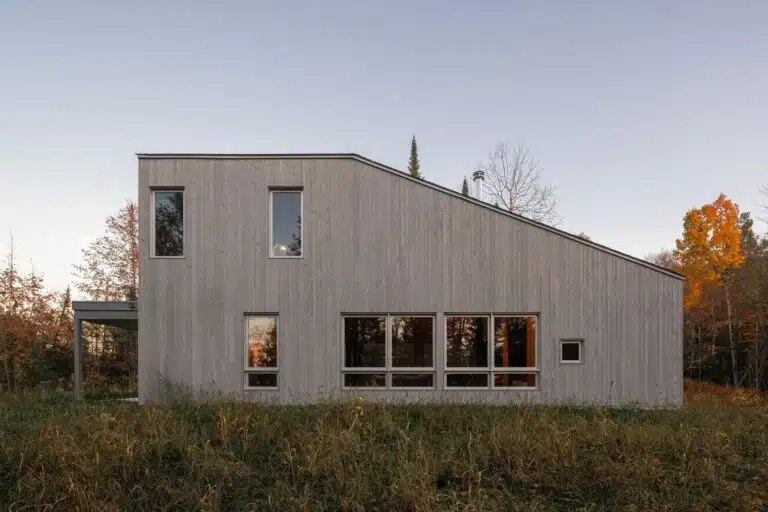 Side view of Scotch Chalet featuring light grey wooden cladding and a sloped roofline amidst a forest in Grenville-sur-la-Rouge.