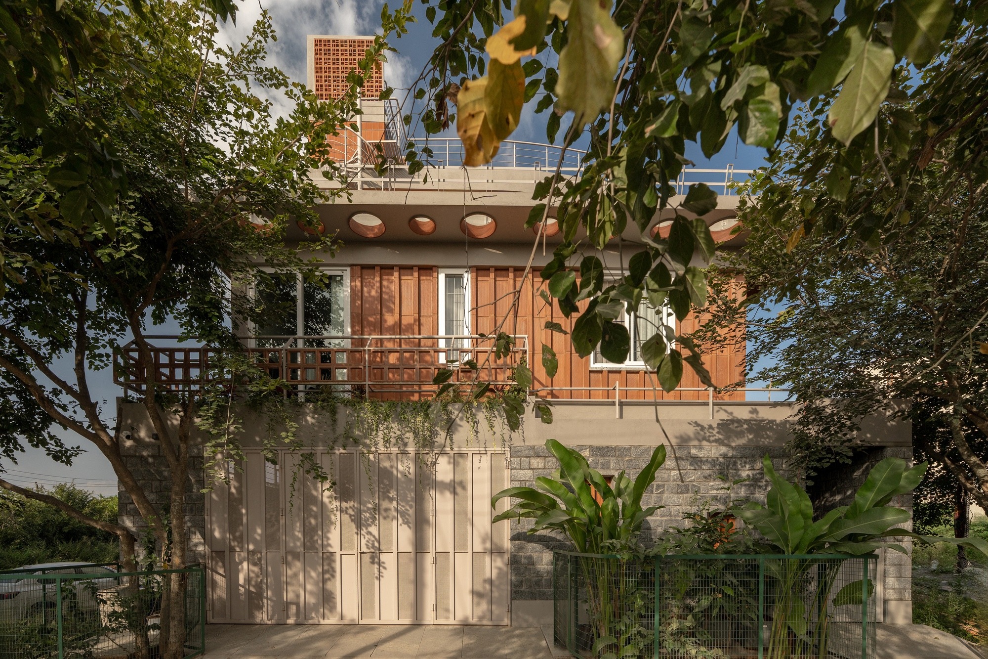 Exterior facade of Thallero House featuring terracotta jali walls, stone masonry at the base, and lush green landscaping.
