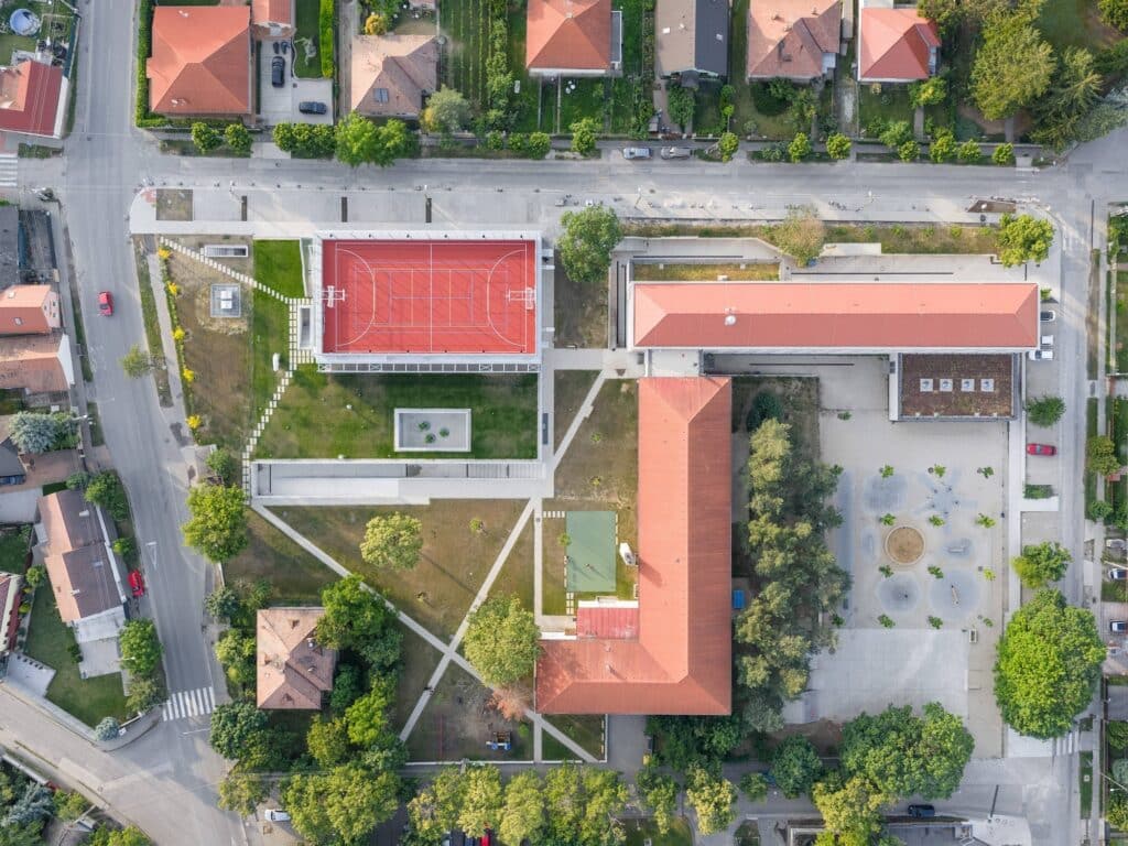 Aerial top-down view of the L-shaped school extension and multipurpose sports hall within the village urban fabric.