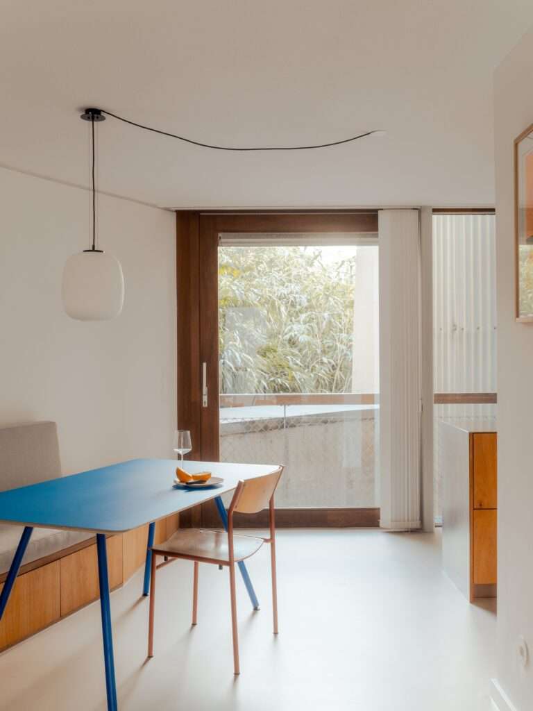 Minimalist dining area in Trommel apartment with a blue table, wooden chair, and a white globe pendant light against a clean white wall.