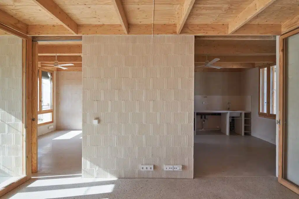 Minimalist interior of a housing unit in Santa Margalida featuring textured white brick walls, exposed wooden beam ceilings, and a simple kitchen counter.
