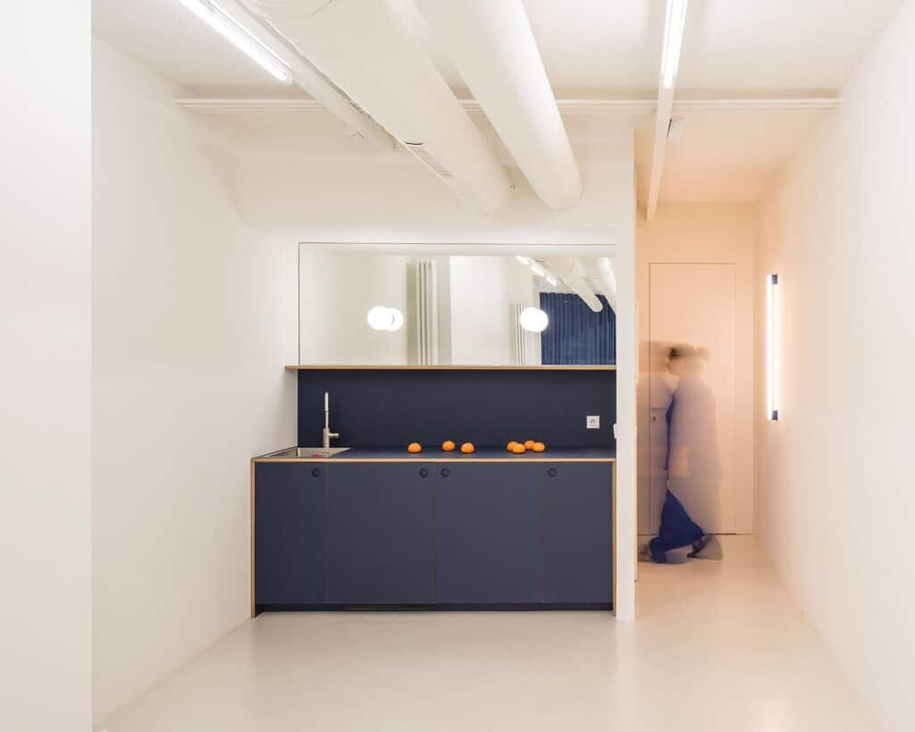 Minimalist interior kitchen area featuring a deep blue counter and cabinetry against neutral white walls with motion blur of a person.