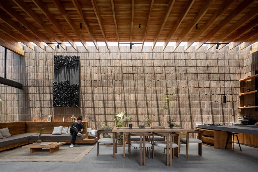 Modern interior living room with exposed brick walls, wooden rafter ceiling, and natural sunlight filtering through high openings.