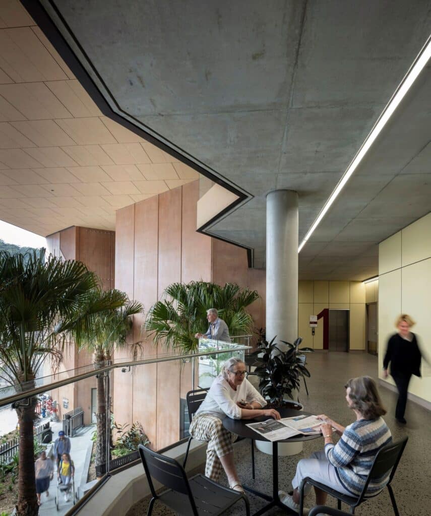 Interior balcony of the Neighborhood Room featuring people at tables, concrete columns, and indoor cabbage tree palms.