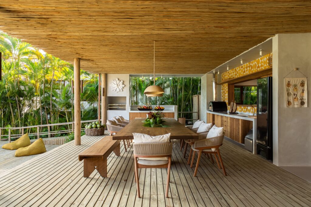 Outdoor dining area with a long wooden table, eucalyptus wood ceiling, and a modern open kitchen under a rustic pergola.