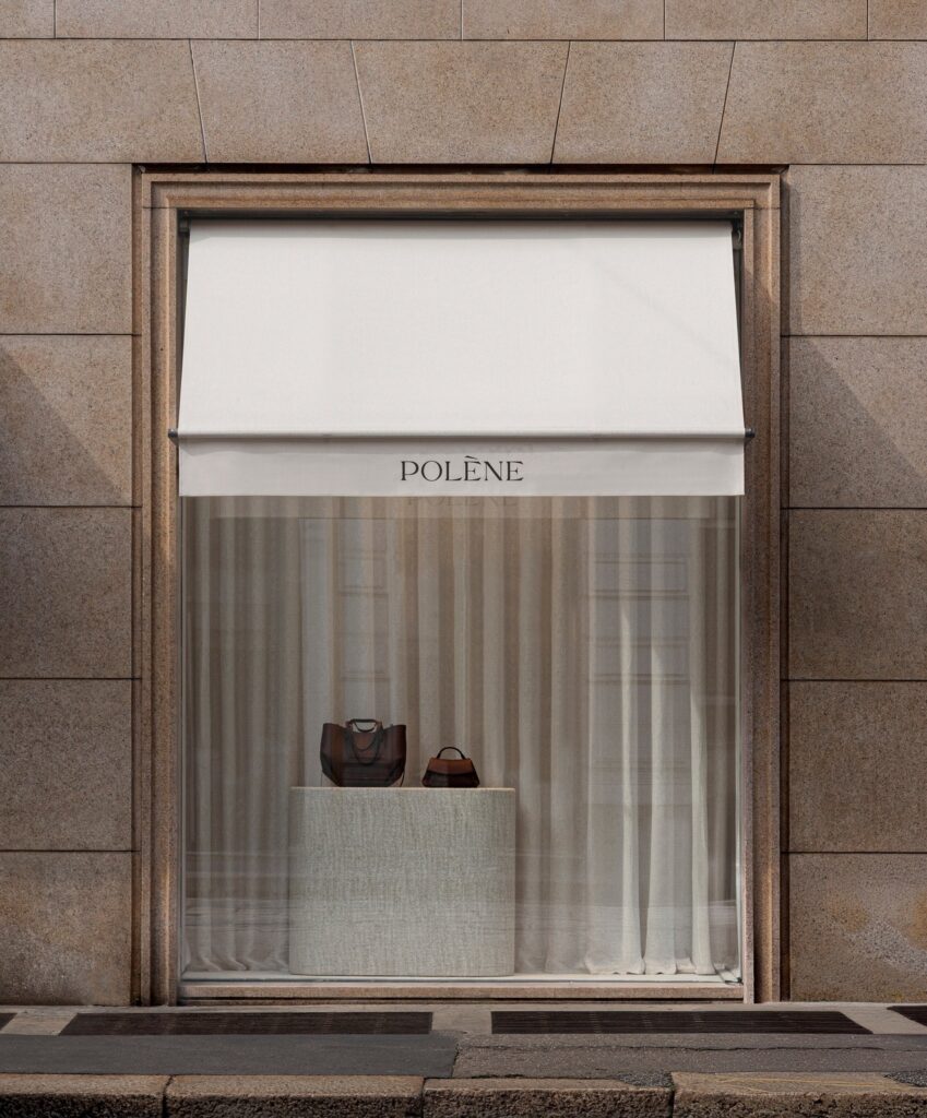 Minimalist storefront window of Pol&egrave;ne Milan featuring a stone plinth with two leather handbags against a backdrop of soft linen curtains and a white awning.