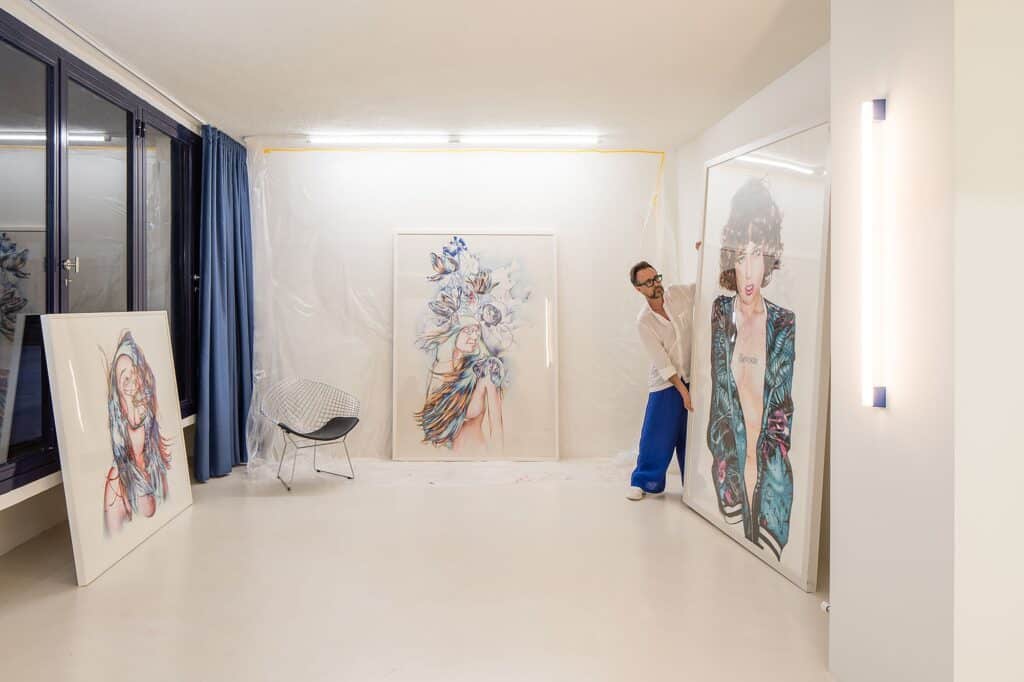 An artist handling a large-scale framed portrait in a bright, high-ceilinged white studio with blue curtains and large windows.