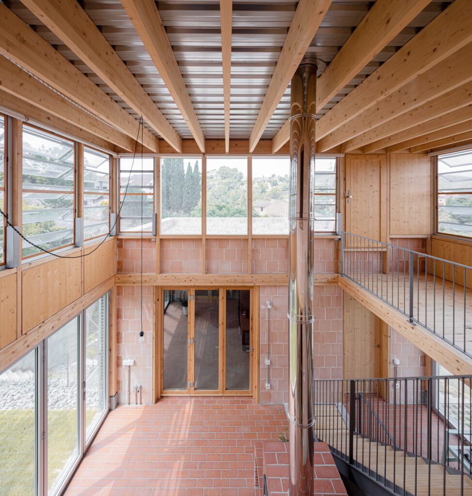 High-angle view of a glass atrium with terracotta tile flooring and a vertical steel chimney pipe under a wooden beam roof.