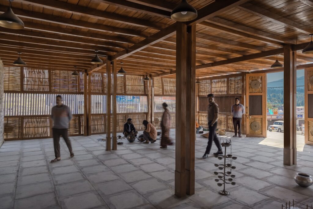 Interior view of a spacious craft workshop with wooden pillars, stone flooring, and artisans working on the ground.