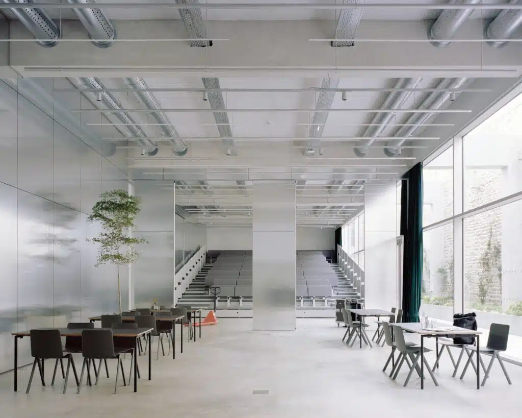 Large multipurpose hall with retractable seating and reflective aluminum wall panels, showcasing flexible interior design.