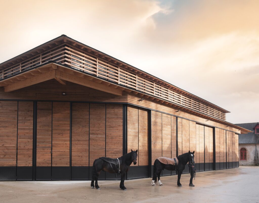Close-up of the timber facade of the Hennebont equestrian hall with two horses in the foreground, highlighting the vertical wooden slats and retracted walls.