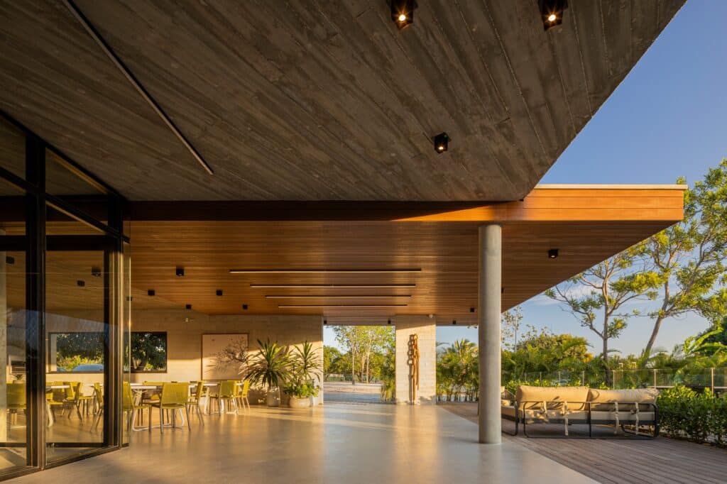 Perspective from under a cantilevered concrete ceiling towards an outdoor lounge area with wooden accents and glass facades.