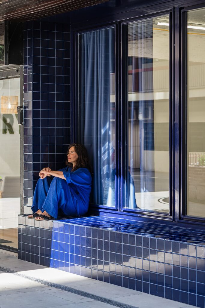 Close-up of a person dressed in blue sitting on the reflective cobalt blue tiled ledge of the building facade.