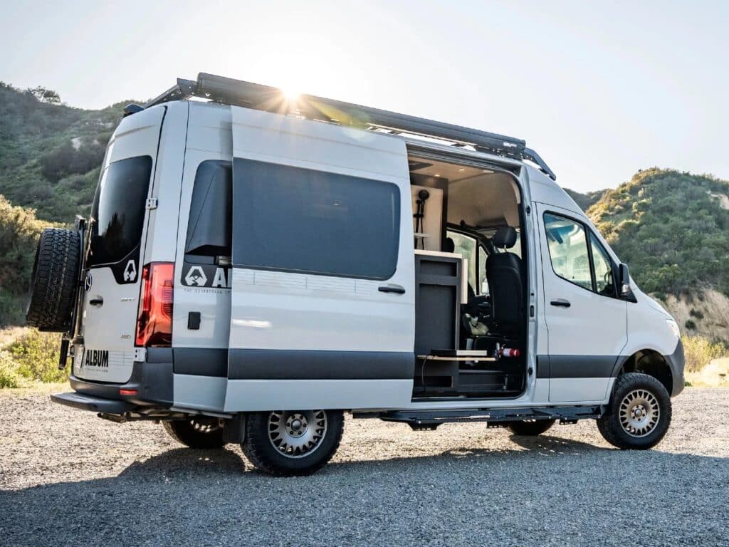 Side view of a silver Mercedes-Benz Sprinter 144 AWD camper van with the sliding door open, showcasing the interior kitchen unit and roof rack.