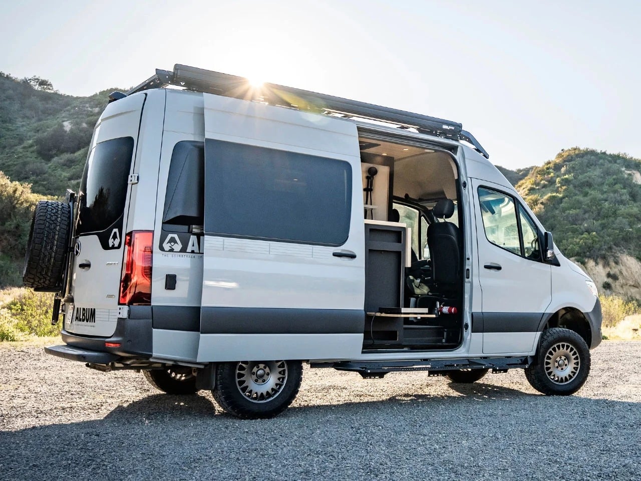Side view of a silver Mercedes-Benz Sprinter 144 AWD camper van with the sliding door open, showcasing the interior kitchen unit and roof rack.