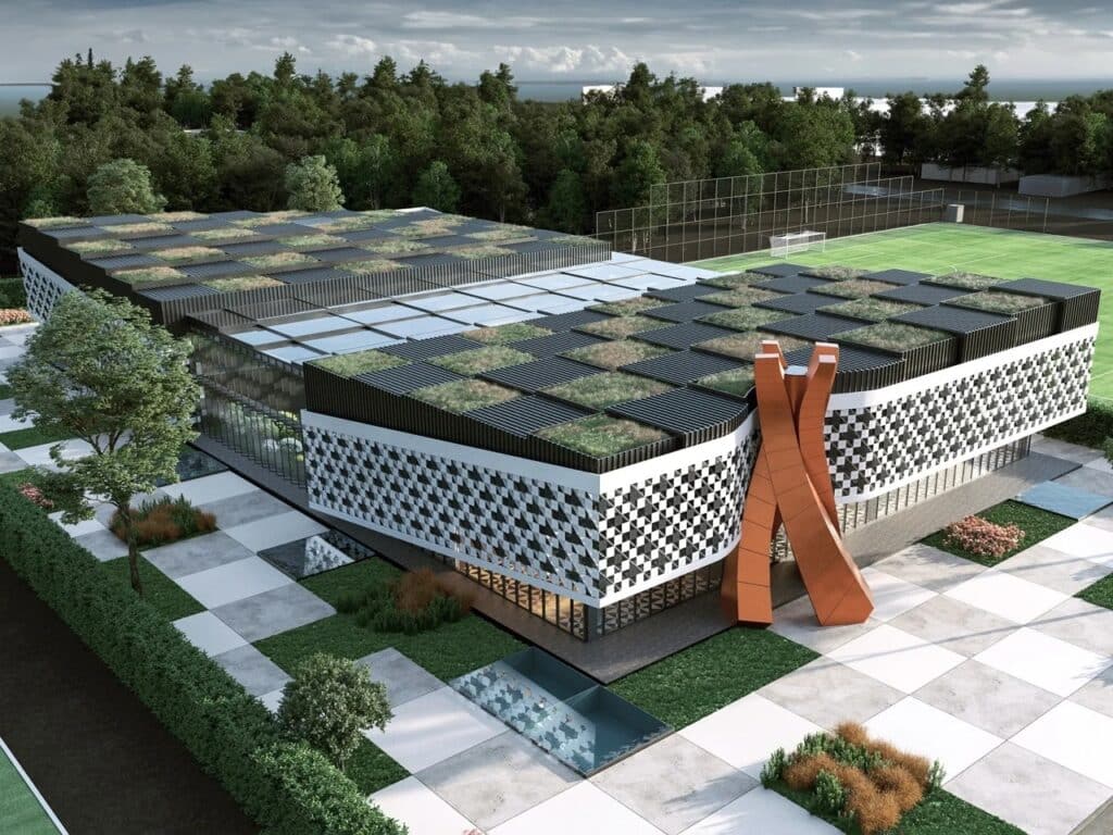 Aerial perspective view of the Batumi Chess Palace building, showing the pixelated black and white facade pattern and the gridded green roof, adjacent to a sports field under a cloudy sky.