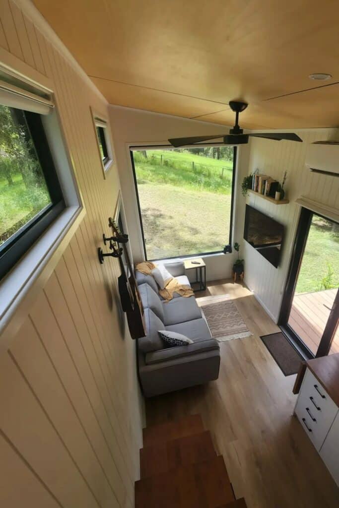 High-angle interior view of a tiny home living room with a grey sofa, large picture window, and plywood ceiling.