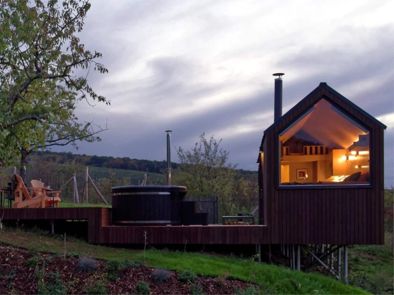 Exterior evening view of NestOff tiny cabin in Hungary, featuring a large gabled window and an outdoor hot tub on a wooden deck.