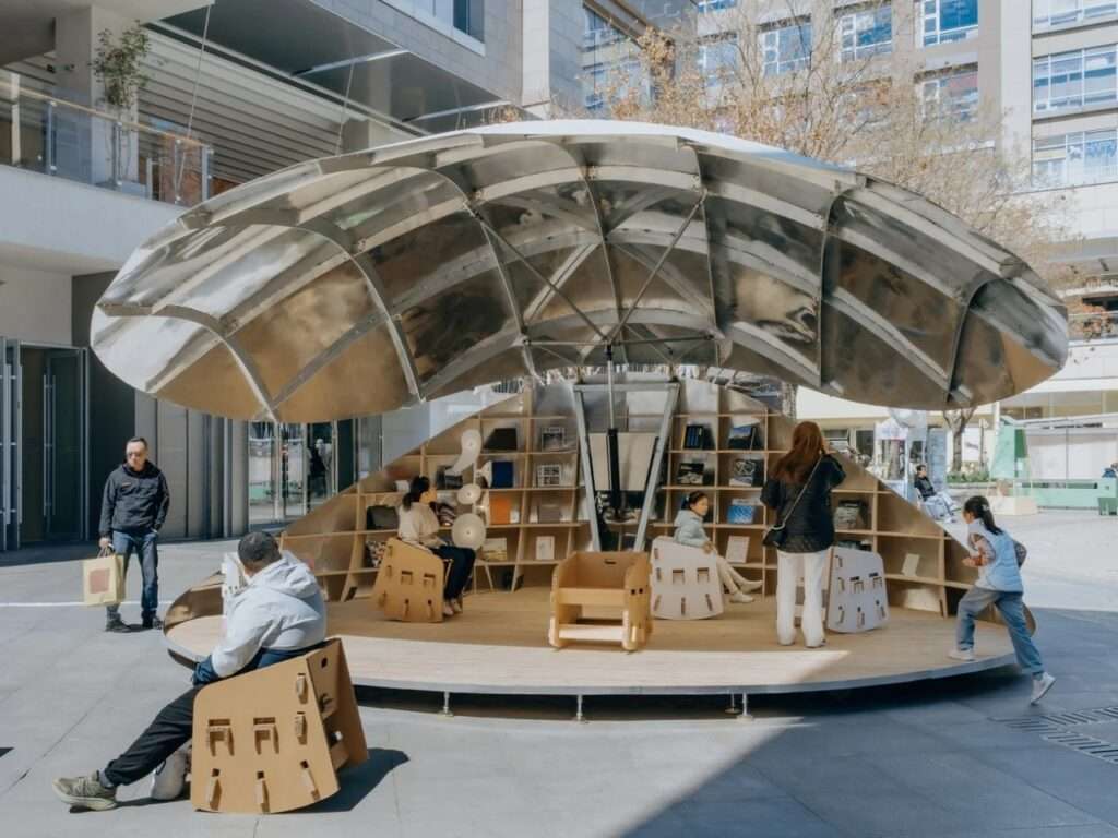 A daytime photograph of the kinetic "Book Shell" pavilion in Beijing, fully opened to form a metallic sun umbrella. Multiple people sit on the wooden deck using wooden-patterned cardboard chairs and browsing bookshelves.