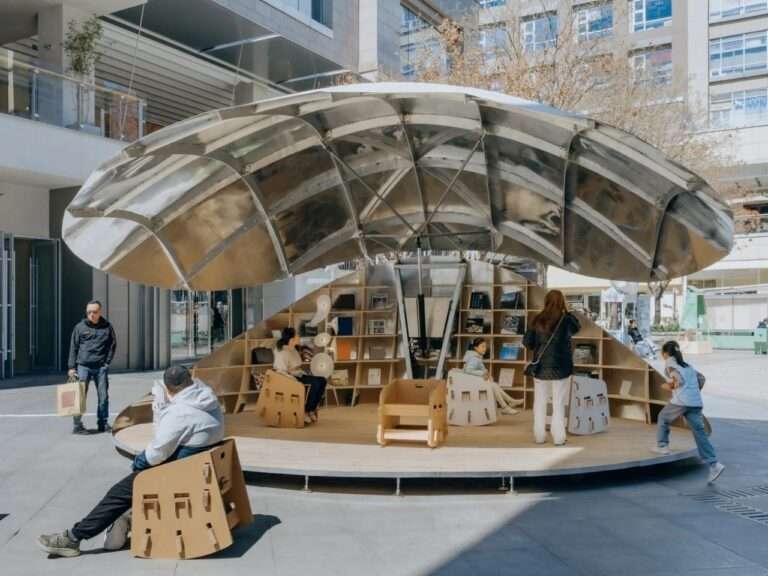 A daytime photograph of the kinetic "Book Shell" pavilion in Beijing, fully opened to form a metallic sun umbrella. Multiple people sit on the wooden deck using wooden-patterned cardboard chairs and browsing bookshelves.