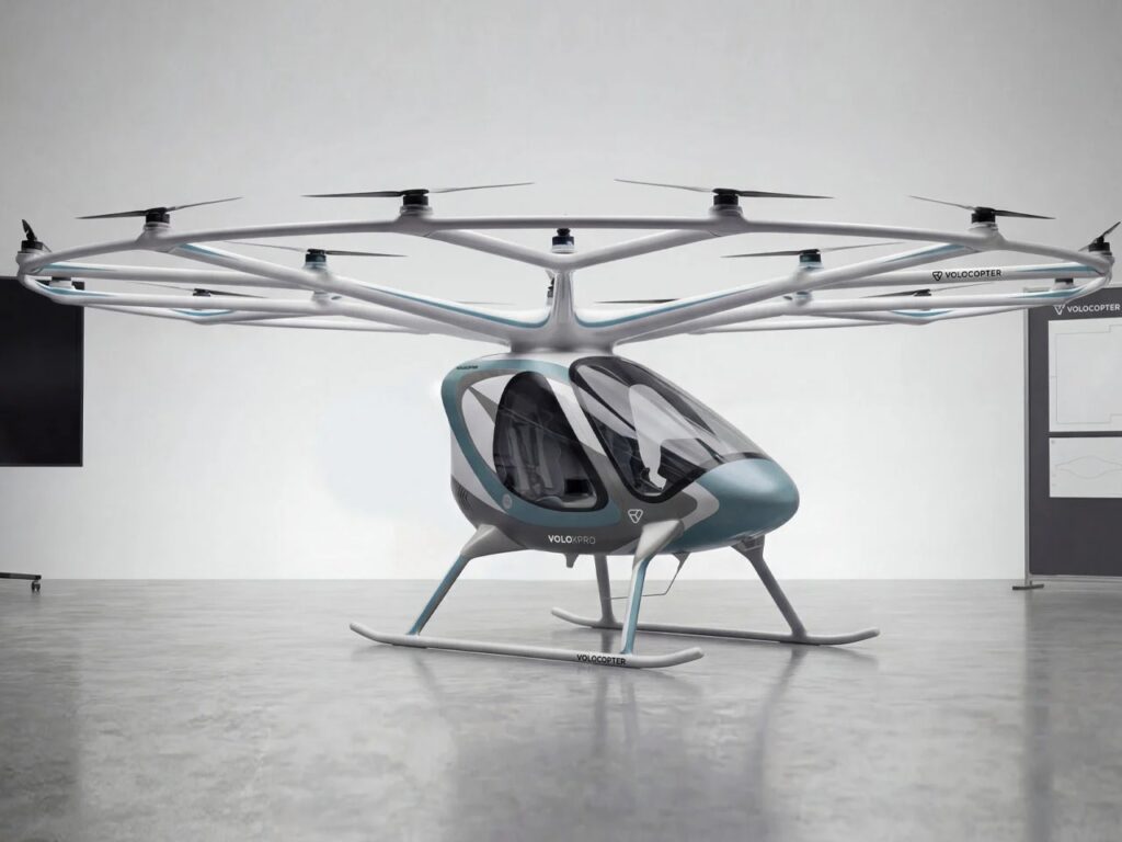 A full side profile view of the white Volocopter VoloXPro eVTOL aircraft with its 18-rotor overhead ring structure, parked on a reflective grey studio floor. The two-seat cabin with its teal-blue nose cone is clearly visible.