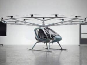A full side profile view of the white Volocopter VoloXPro eVTOL aircraft with its 18-rotor overhead ring structure, parked on a reflective grey studio floor. The two-seat cabin with its teal-blue nose cone is clearly visible.
