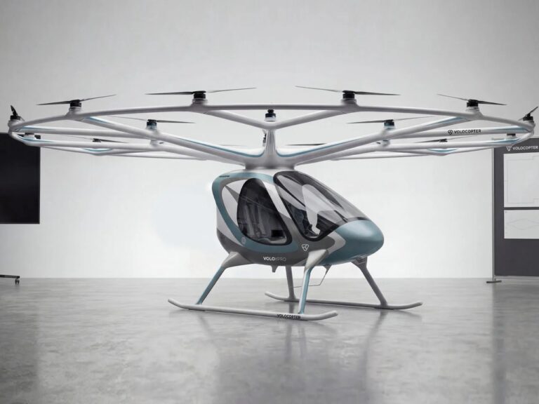A full side profile view of the white Volocopter VoloXPro eVTOL aircraft with its 18-rotor overhead ring structure, parked on a reflective grey studio floor. The two-seat cabin with its teal-blue nose cone is clearly visible.