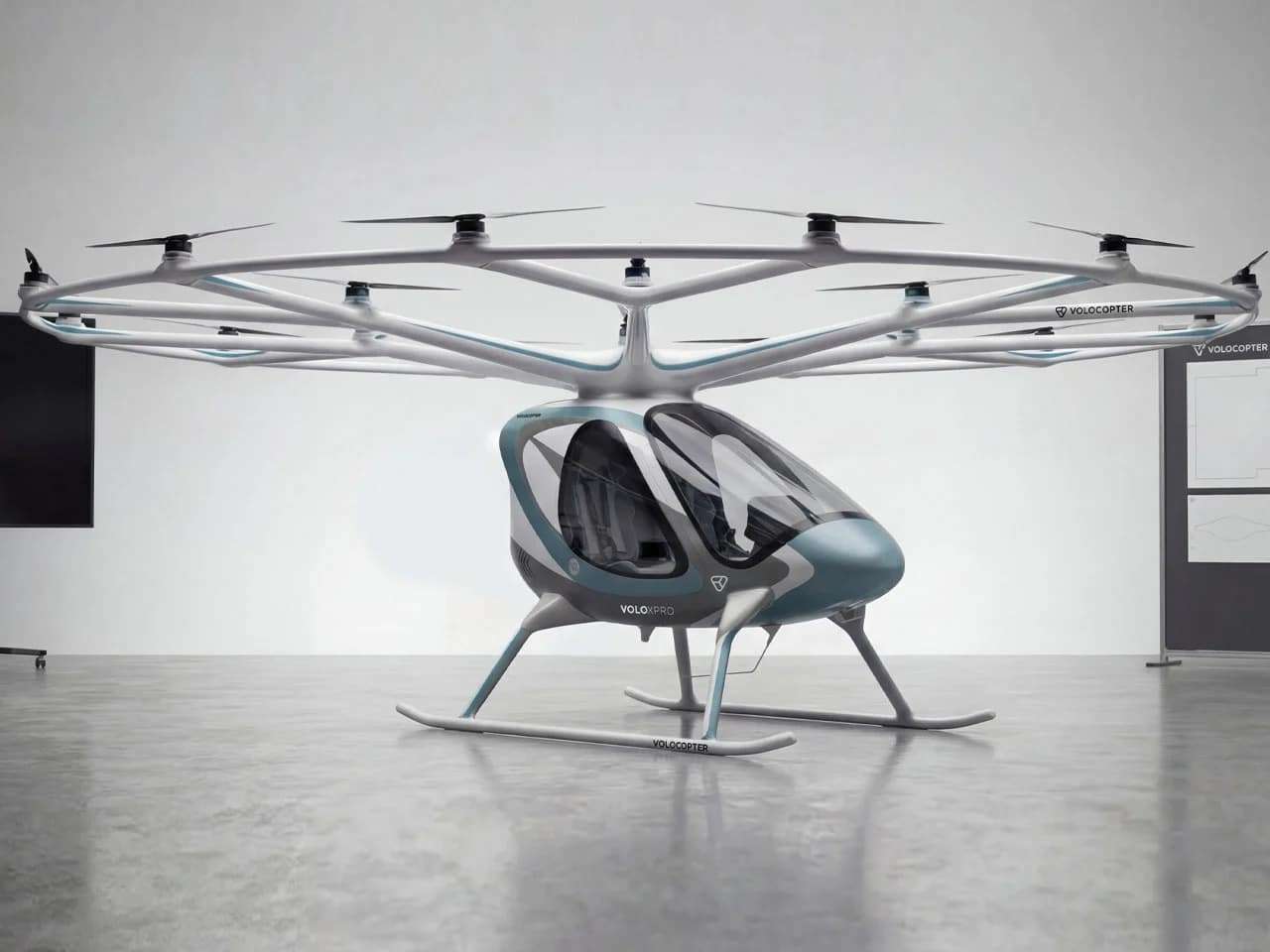 A full side profile view of the white Volocopter VoloXPro eVTOL aircraft with its 18-rotor overhead ring structure, parked on a reflective grey studio floor. The two-seat cabin with its teal-blue nose cone is clearly visible.