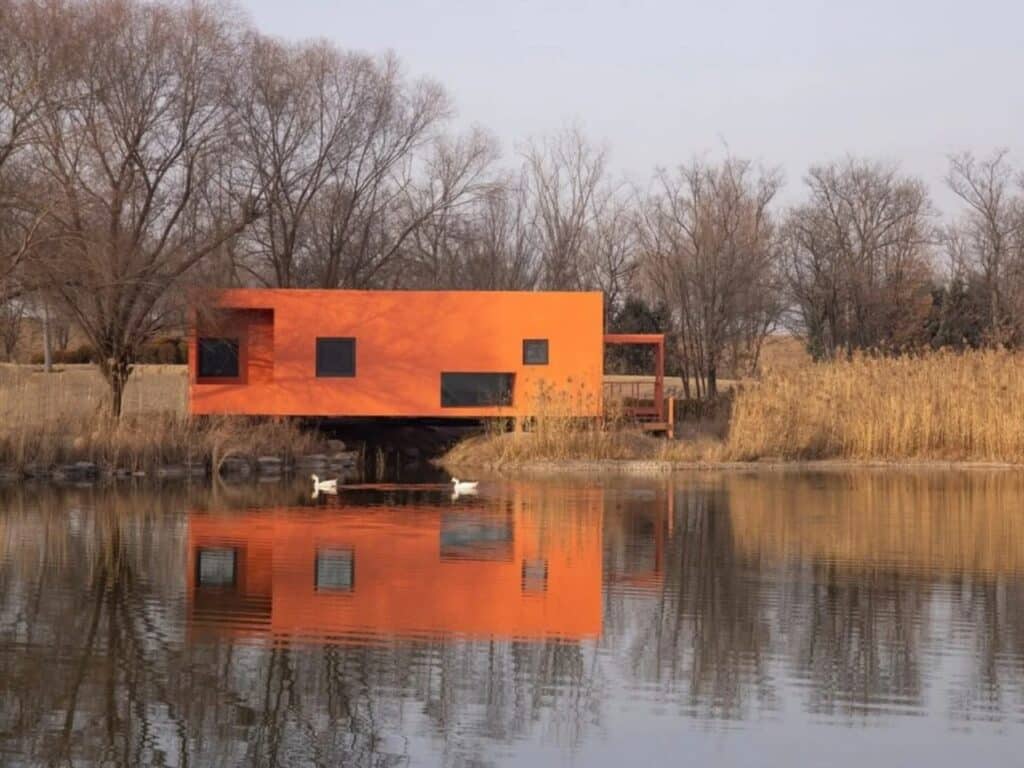 A horizontal wide shot of the Red Bridge Cabin by Wiki World, a minimalist red timber structure perfectly reflected in a calm lake in Zhengzhou, China.